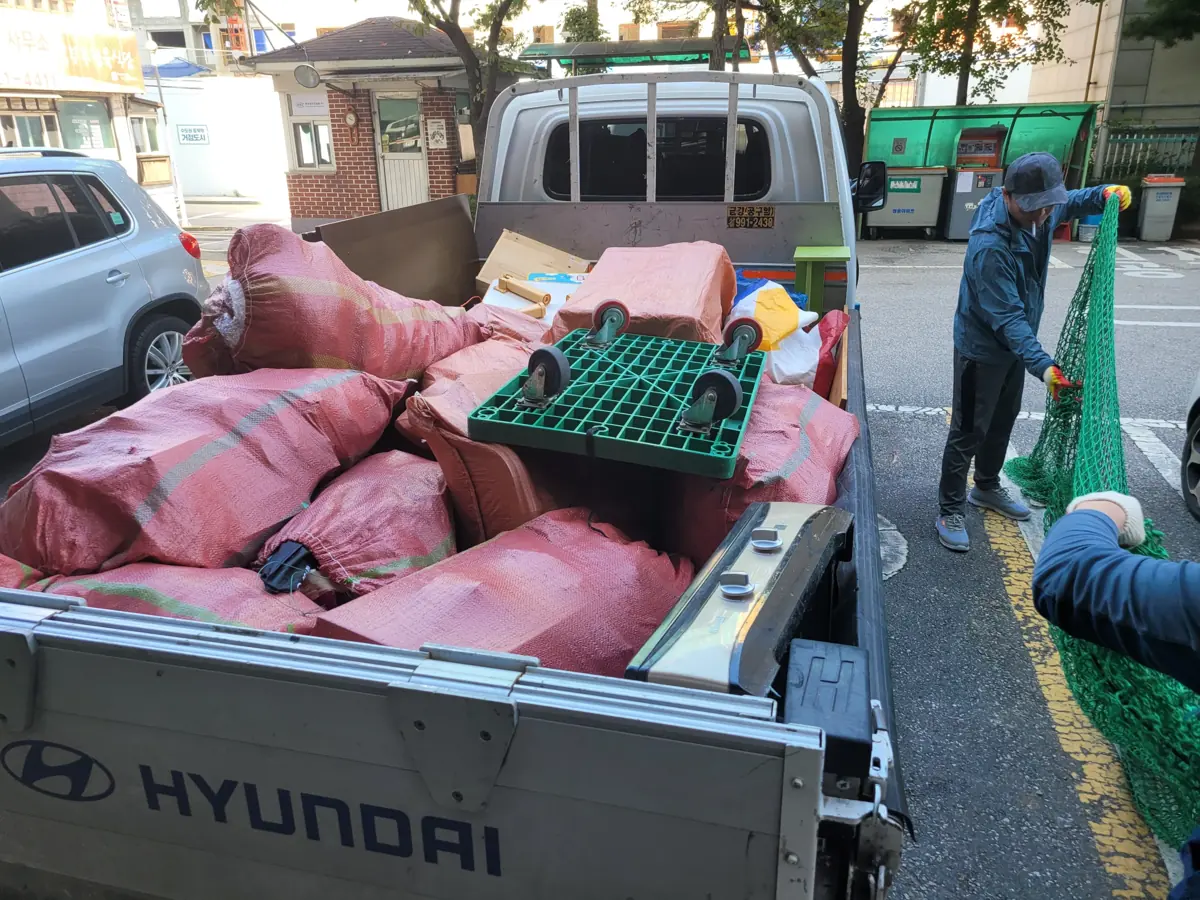 좁은 도로변에 세운 작업 차량 적재함에 자루를 쌓고 작업자가 망을 씌우는 차량 접근 위치