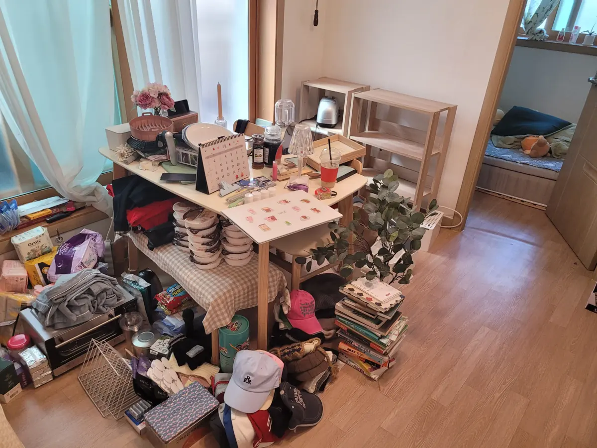 작은 거실 테이블 위와 바닥에 생활물품이 가득 쌓여 집 전체 정리 상담이 필요한 현장